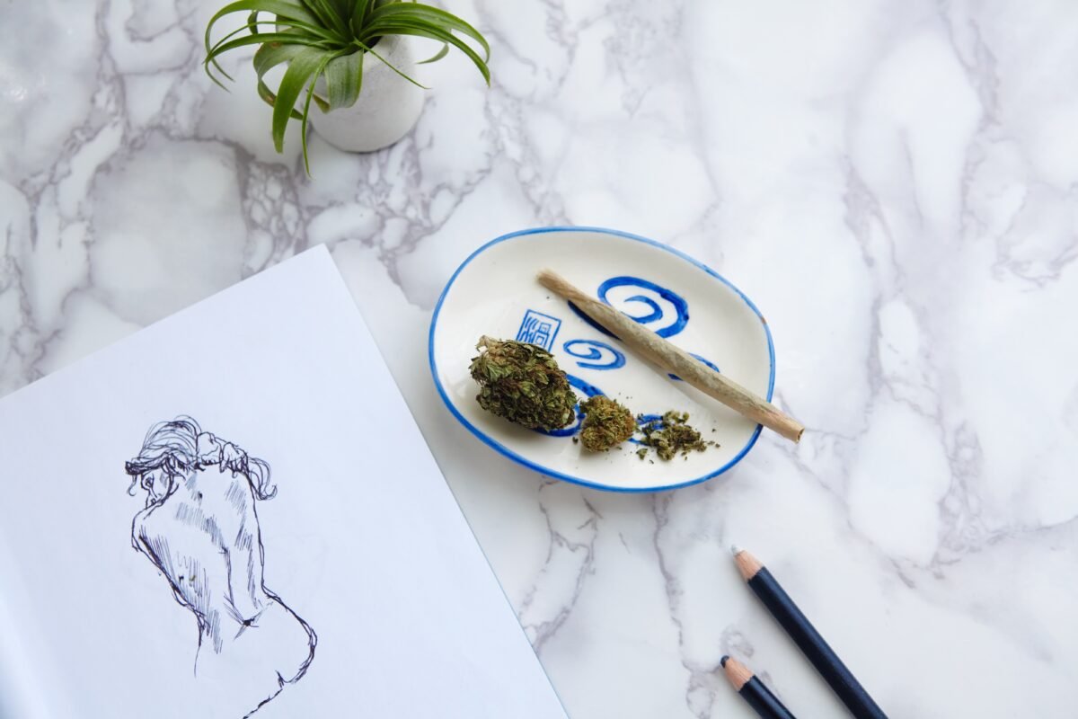 THC CBD Marijuana joint and flowers on ash tray with nude illustration on drawing pad on marble table and succulents.